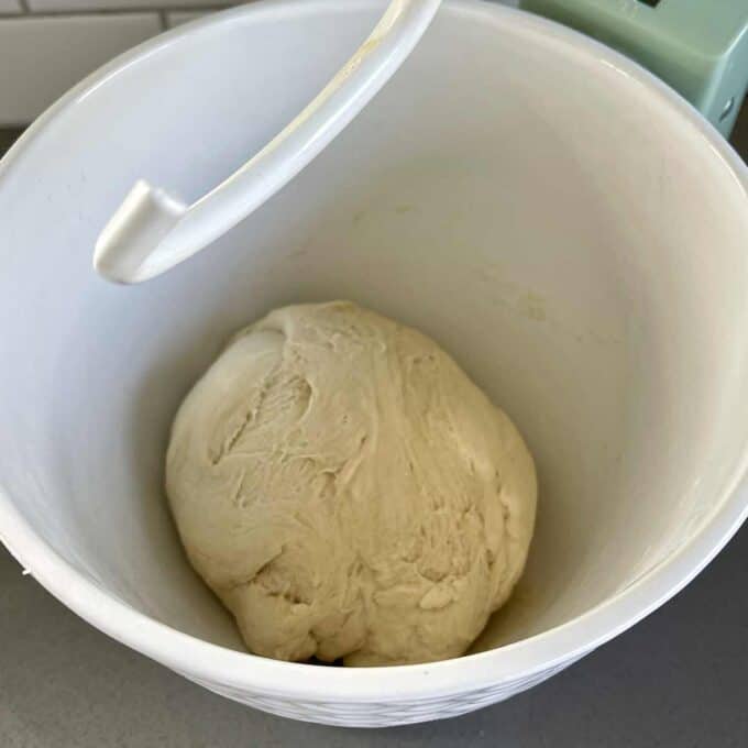 A ball of bread dough rests in a white mixing bowl with a dough hook.