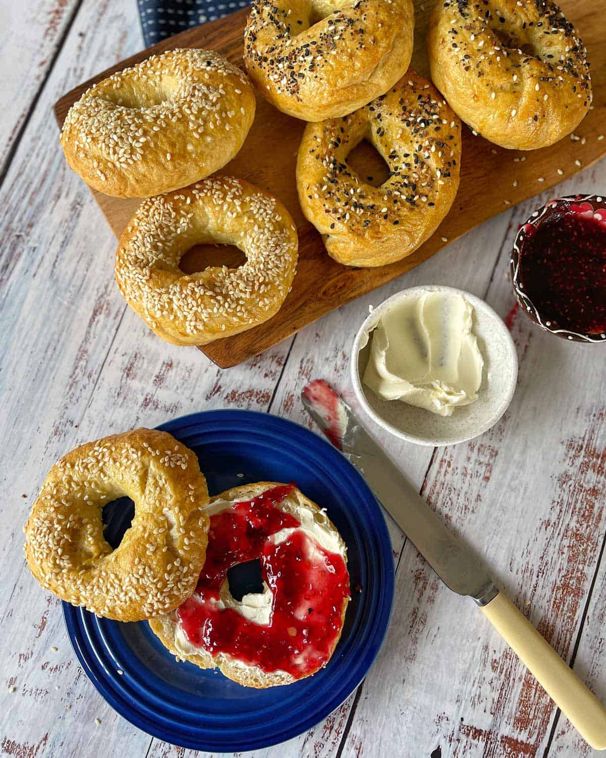 Homemade Bagels Bagels topped with sesame and poppy seeds on a wooden board. One bagel on a blue plate spread with cream cheese and red jam. Knife and bowl of cream cheese nearby. Rustic white table surface.