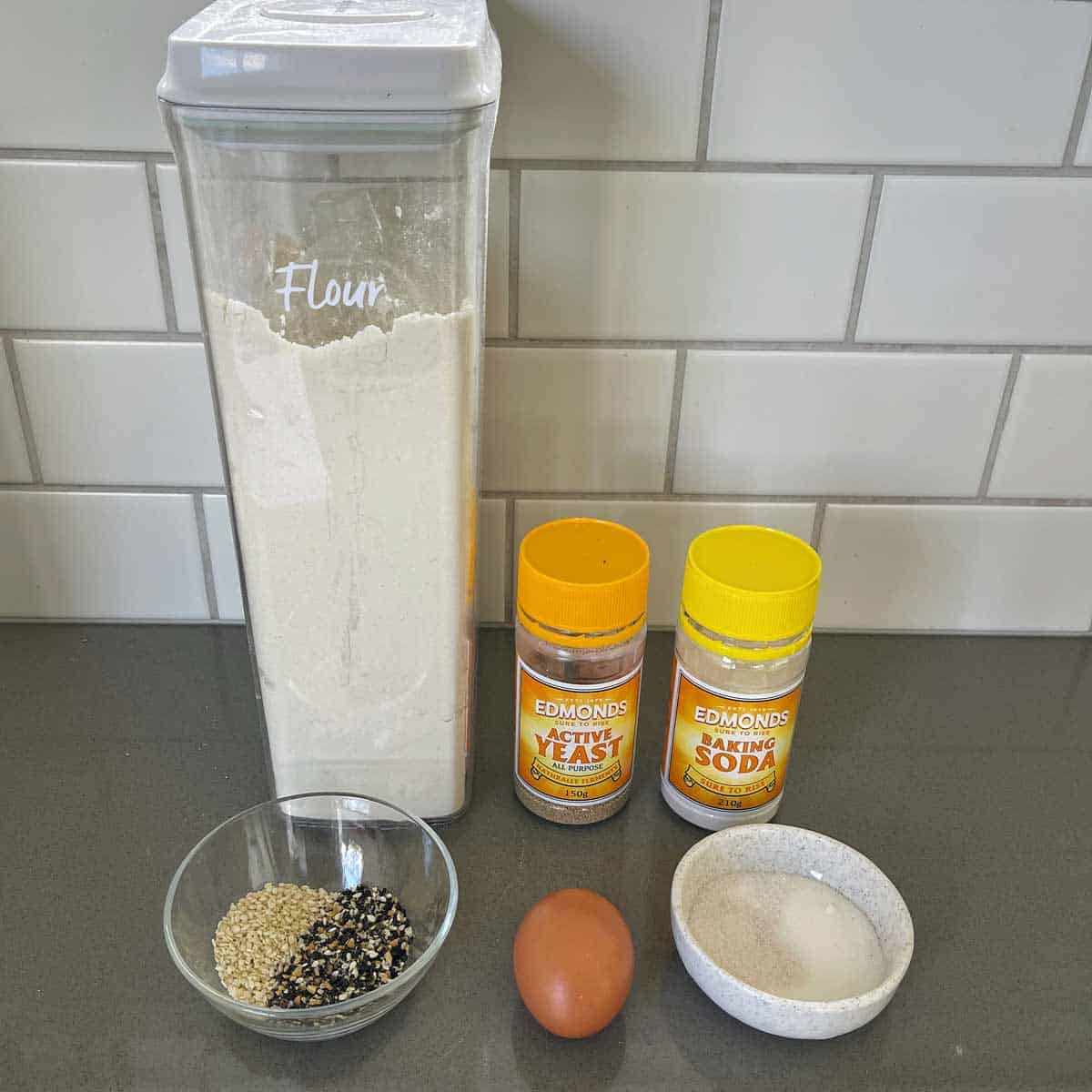 Flour container, jars of yeast and baking soda, a bowl of mixed seeds, an egg, and a bowl of sugar on a kitchen counter with a tiled backsplash.