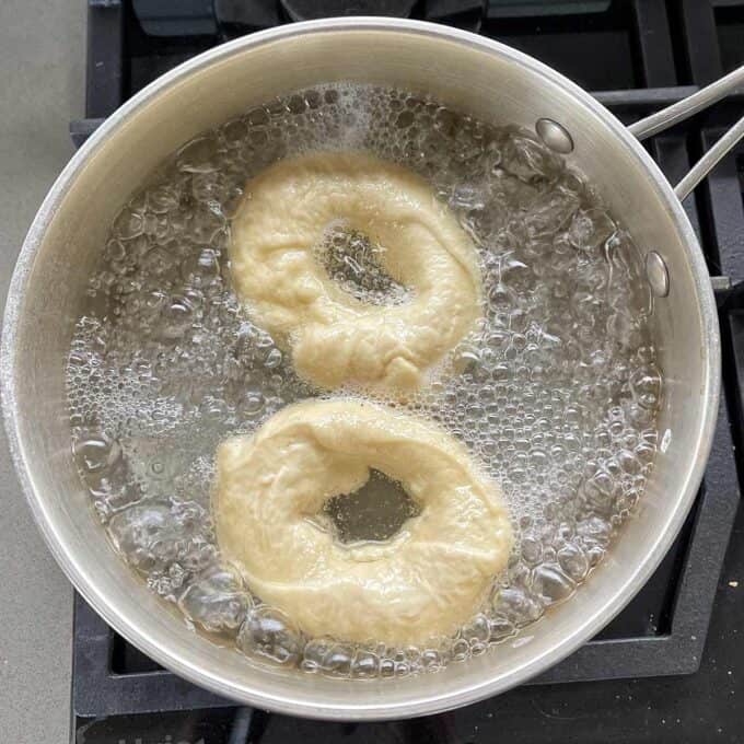 Two bagels boiling in a metal pot on a stove.