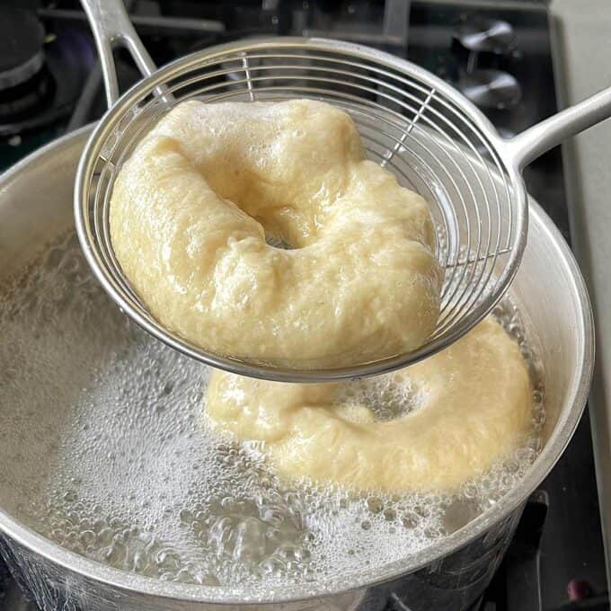 Bagel dough being boiled with a slotted spoon lifting one from the pot.