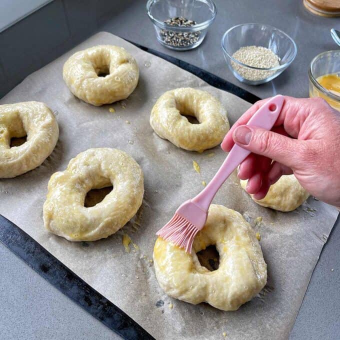 A hand uses a pink brush to apply egg wash to unbaked bagels on parchment paper. In the background, there are small bowls with seeds.