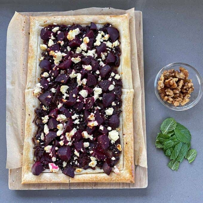 A beetroot and feta tart on a chopping board sitting next to a bowl of walnuts and a bunch of french mint leaves.
