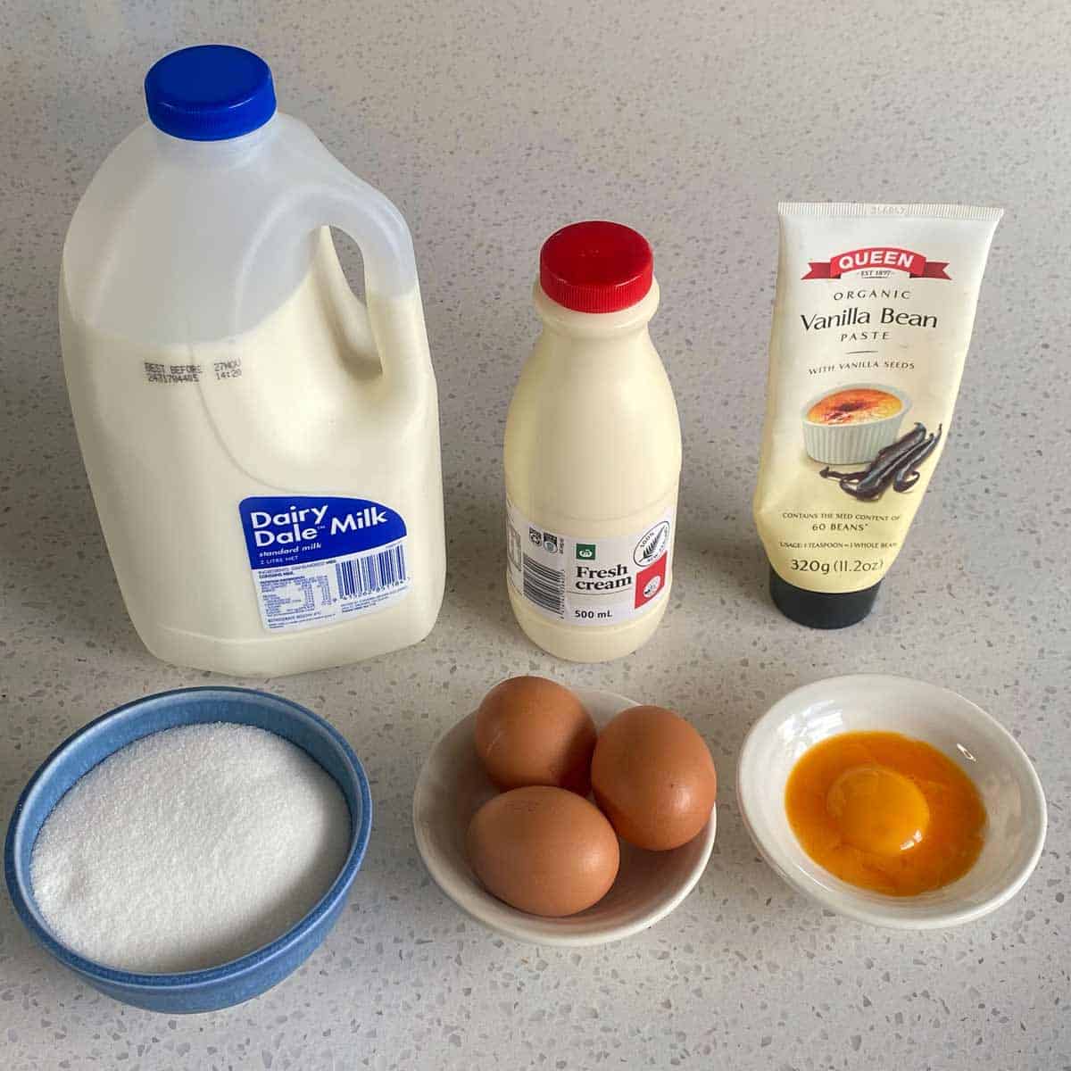 Milk, fresh cream, vanilla bean paste, sugar, three whole eggs, and one egg yolk on a countertop.