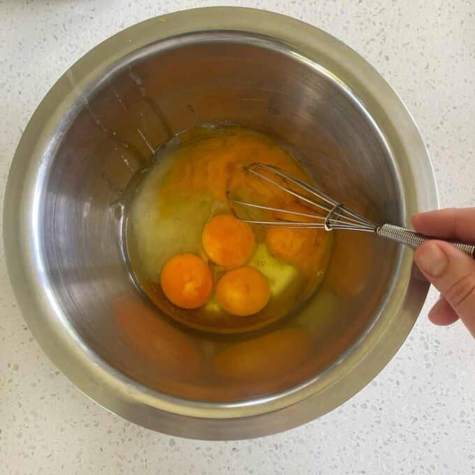 A hand holds a whisk over a metal bowl containing three cracked eggs on a white countertop.