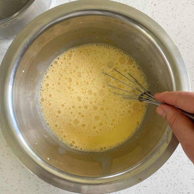 A hand whisking a yellow liquid mixture in a stainless steel bowl, with a white countertop in the background.