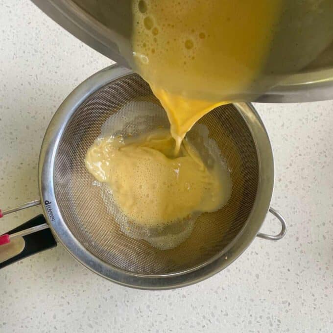 Egg mixture being poured through a metal strainer into a bowl on a white countertop.