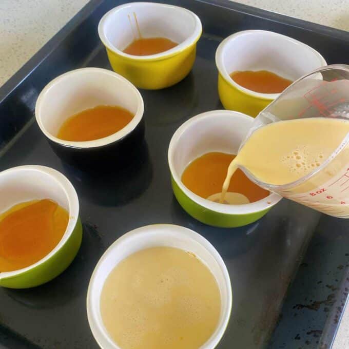 Creamy mixture being poured into ramekins with caramel sauce on a baking tray, preparing for baking.