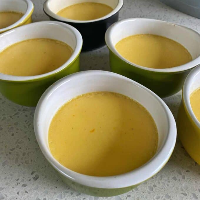 Six ramekins filled with smooth yellow custard are arranged on a speckled countertop.