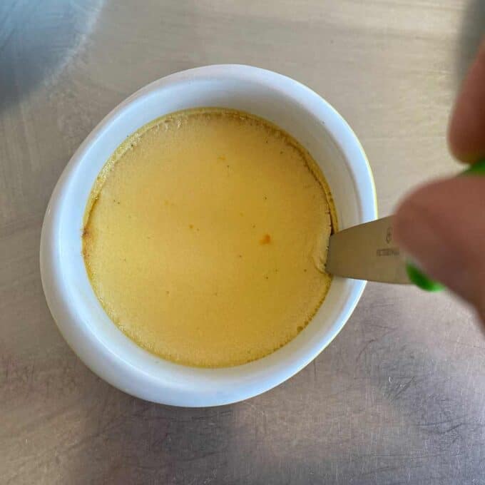 A person inserts a knife into a small dish of yellow custard, set on a metal surface.