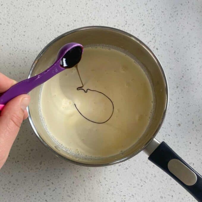 Hand adding vanilla extract with a purple spoon to a saucepan of creamy liquid on a speckled countertop.