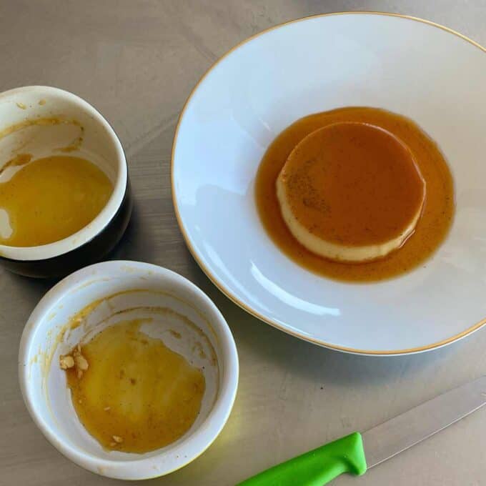 A flan dessert with caramel sauce on a white plate next to two empty ramekins and a green-handled knife on a metal surface.