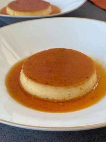 A round flan dessert with caramel sauce on top is placed on a white plate with a grey background.