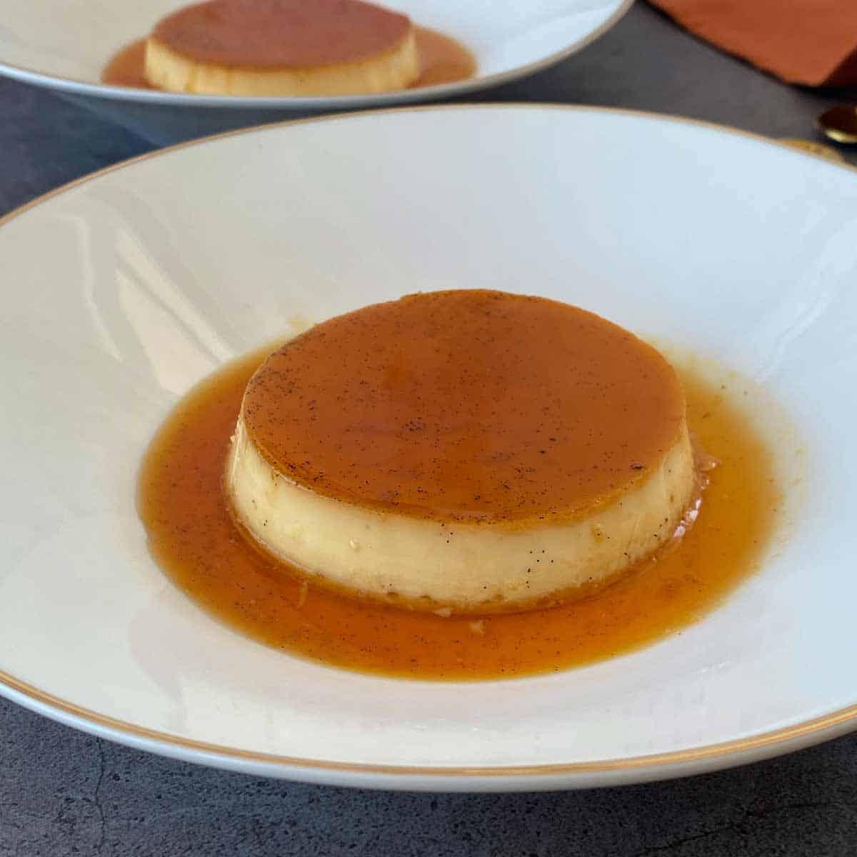 A round flan dessert with caramel sauce on top is placed on a white plate with a grey background.