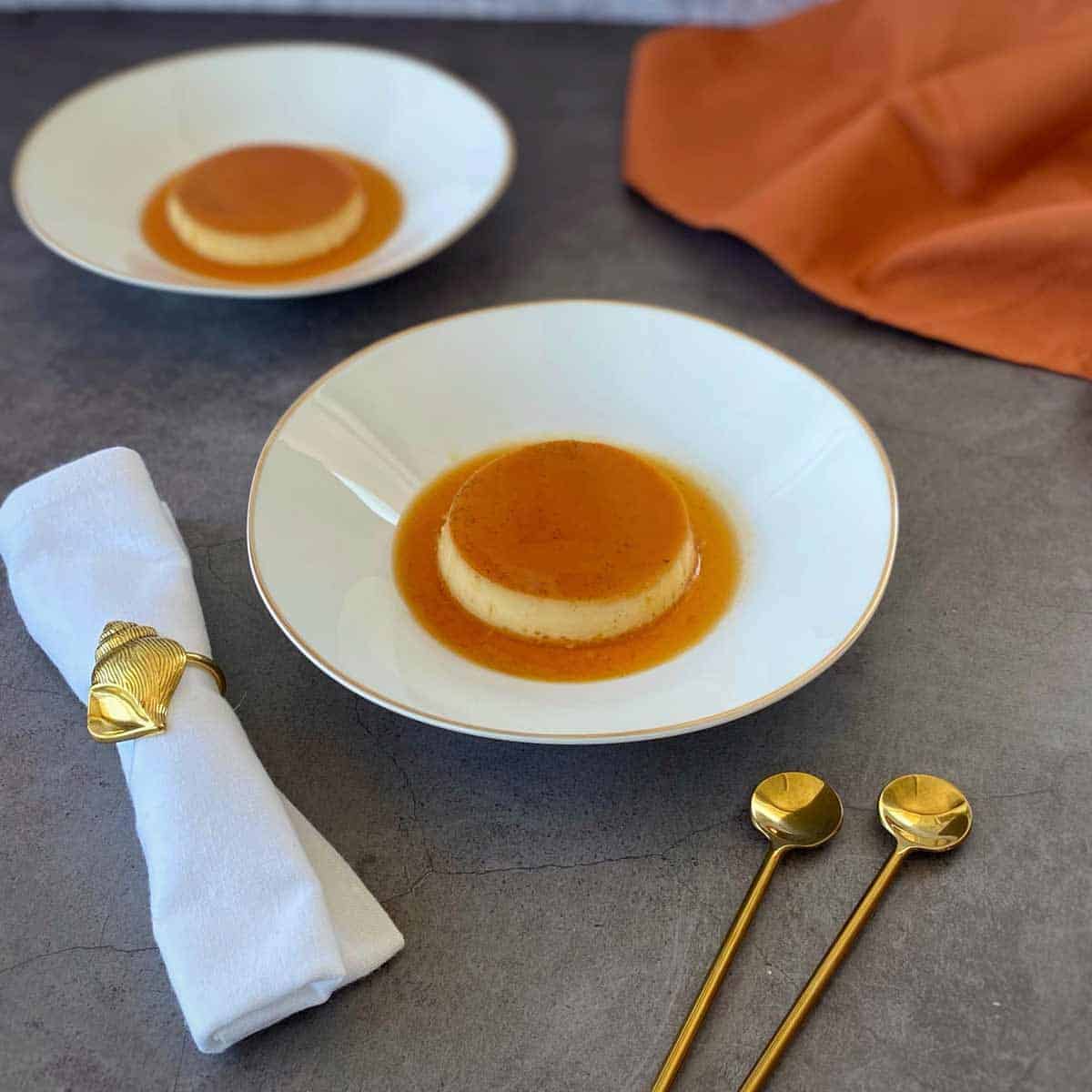 Two plates of flan with caramel sauce on a gray surface, accompanied by a folded napkin with gold ring and two gold spoons. An orange napkin is in the background.