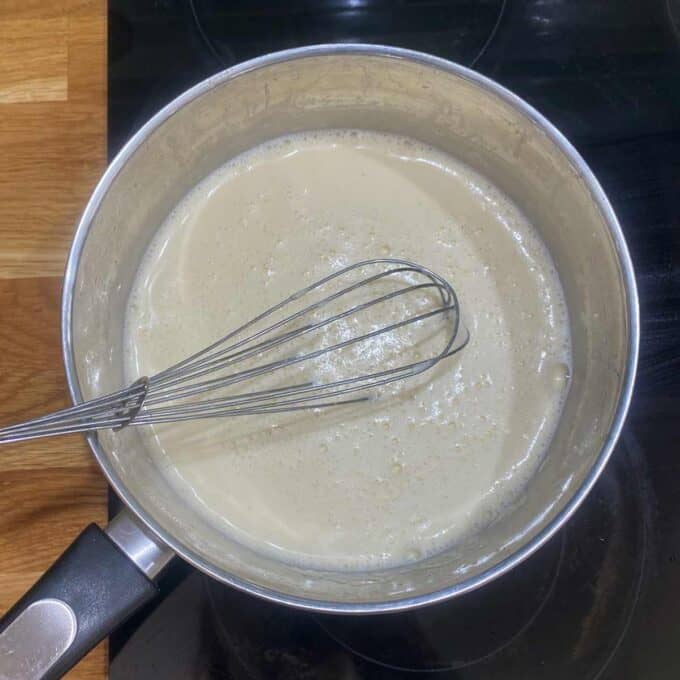 A saucepan on a stove contains a light beige liquid being stirred with a metal whisk.