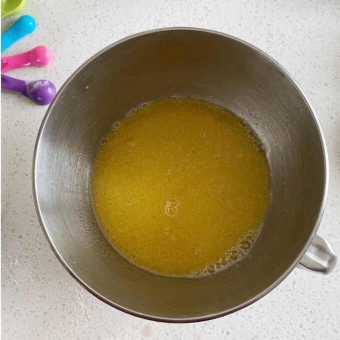 Stainless steel mixing bowl containing a yellow liquid mixture on a white countertop, with colorful measuring spoons in the background.