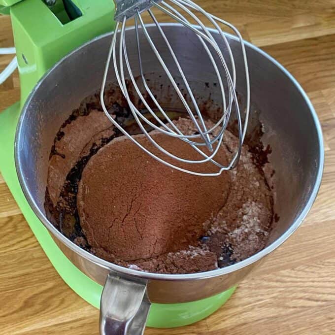 A green stand mixer bowl with cocoa powder and other ingredients being mixed with a wire whisk attachment.
