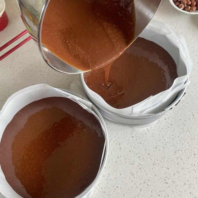 Pouring chocolate cake batter into two round baking pans lined with parchment paper on a countertop.