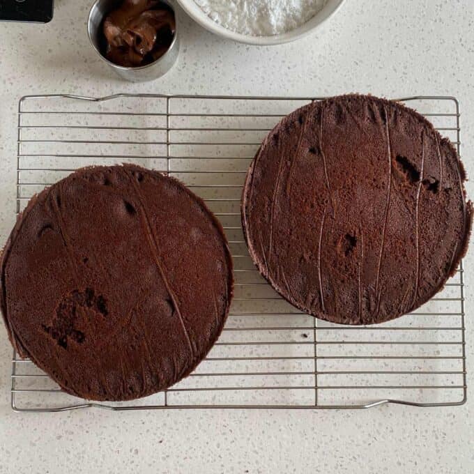 Two round chocolate cakes cooling on a wire rack, with a bowl of powdered sugar and a small cup of chocolate spread nearby.