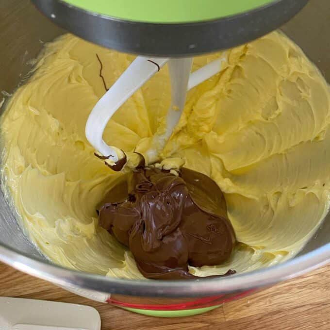 Butter and melted chocolate are in a mixing bowl, being blended with a stand mixer on a wooden countertop.