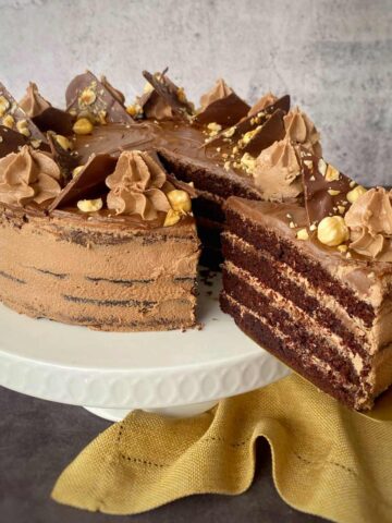 A sliced chocolate cake with layered frosting is displayed on a white cake stand. Topped with chocolate pieces, cream swirls, and nuts. A yellow cloth is draped at the base.