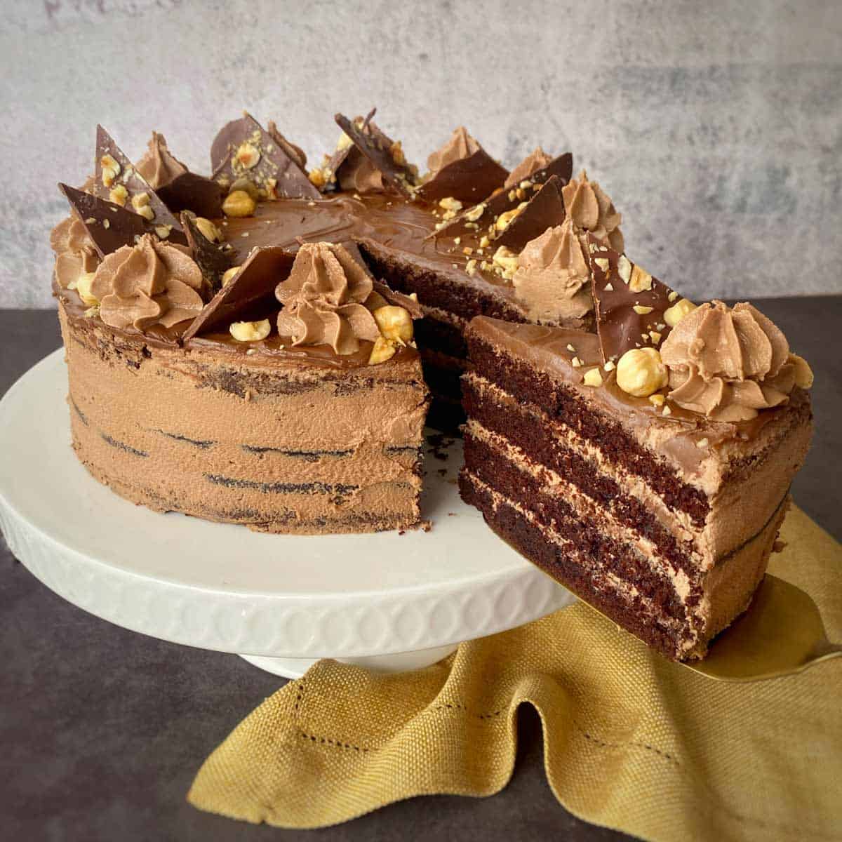 A sliced chocolate cake with layered frosting is displayed on a white cake stand. Topped with chocolate pieces, cream swirls, and nuts. A yellow cloth is draped at the base.