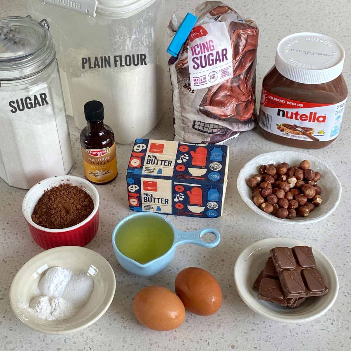 Assorted baking ingredients on a counter include flour, sugar, cocoa, butter, eggs, Nutella, icing sugar, vanilla extract, nuts, chocolate, and baking powder.