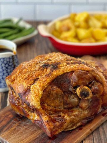 Roast pork with crispy skin on a wooden board, sauce in a small jug, green beans, and roasted potatoes in a dish in the background.