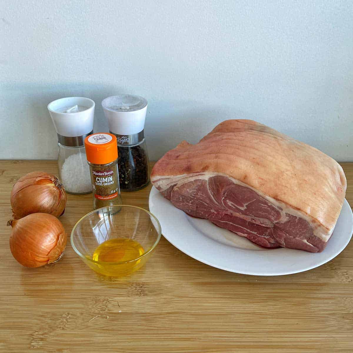 A raw pork shoulder on a plate with onions, a bowl of oil, cumin seasoning, and salt and pepper grinders on a wooden surface.