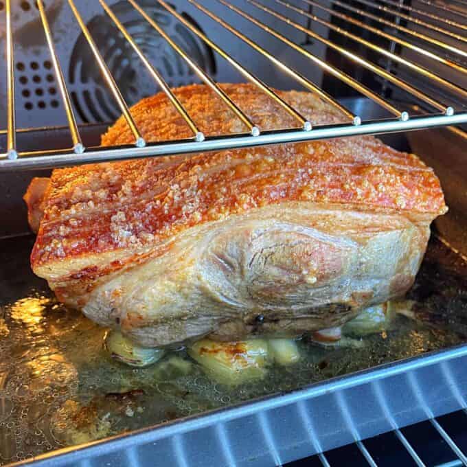 A large piece of pork with crispy skin is roasting in an oven. Drippings collect at the bottom of the pan.