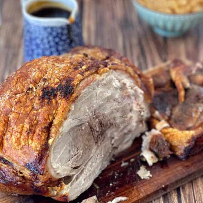 Roast pork with crispy crackling on a wooden board, accompanied by a blue jug of sauce and a bowl of dip in the background.