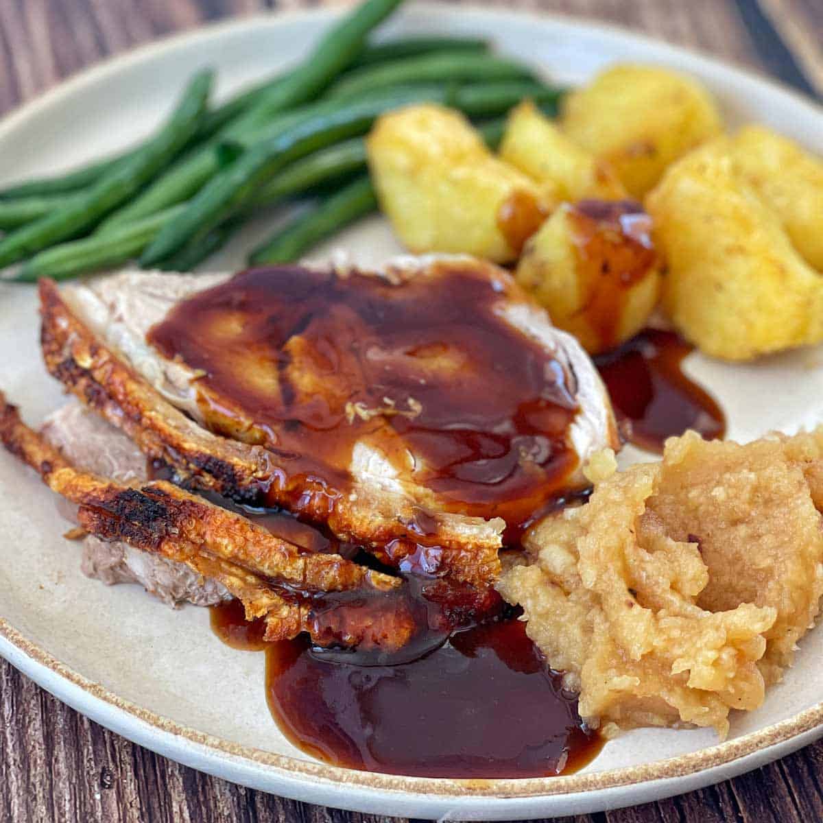 Plate with sliced roast meat covered in gravy, accompanied by green beans, roast potatoes, and applesauce.