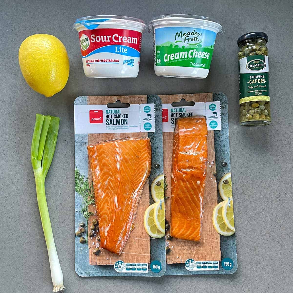 Smoked salmon packs, sour cream, cream cheese, sliced spring onion, a lemon, and a jar of capers arranged on a gray surface.
