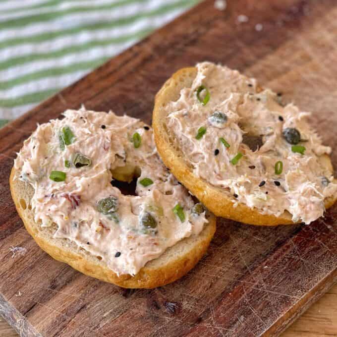 Bagel topped with a creamy spread, capers, and chopped green onions on a wooden board.