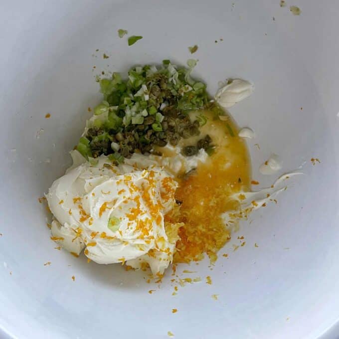 A white bowl containing cream cheese, chopped green onions, capers, minced herbs, orange zest, and a small amount of liquid.