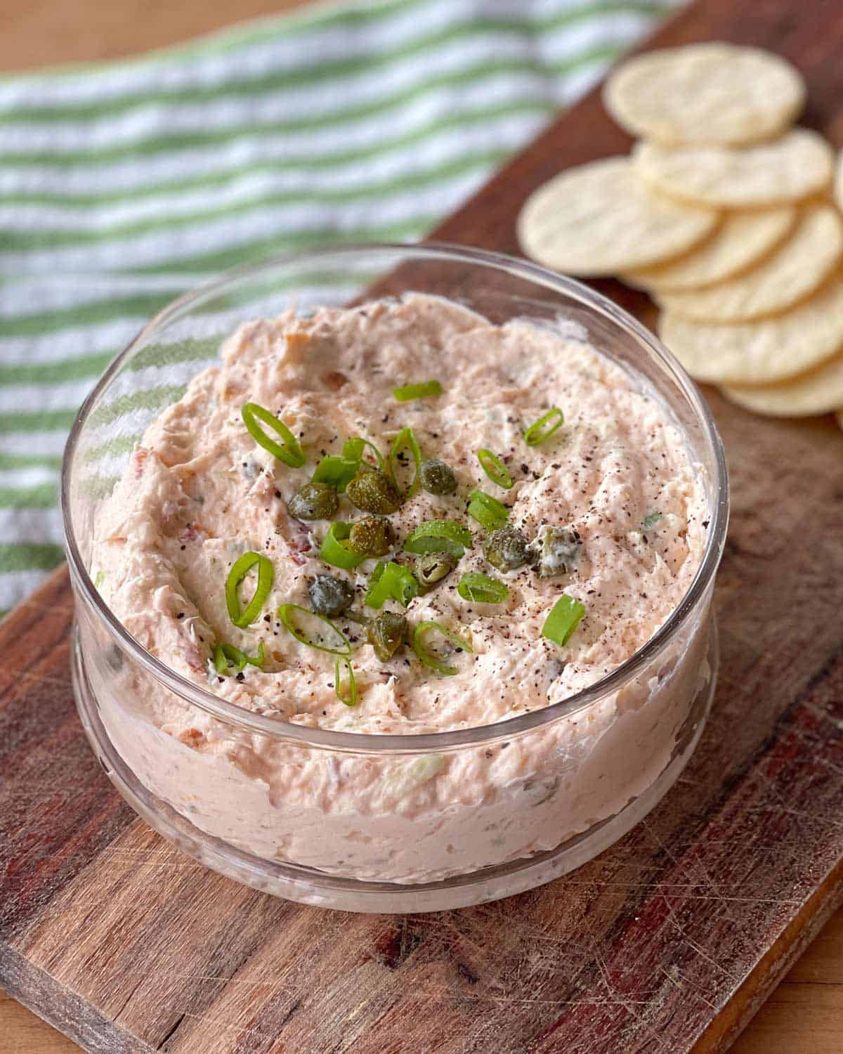 Smoked Salmon Dip A glass bowl of creamy dip topped with chopped green onions and capers sits on a wooden board. Round crackers are next to the bowl.