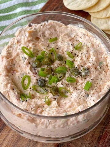A bowl of creamy dip topped with chopped green onions and capers, set on a wooden board. Slices of bread or crackers are partially visible in the background.