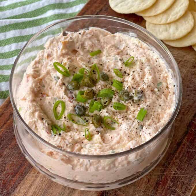 A bowl of creamy dip topped with chopped green onions and capers, set on a wooden board. Slices of bread or crackers are partially visible in the background.