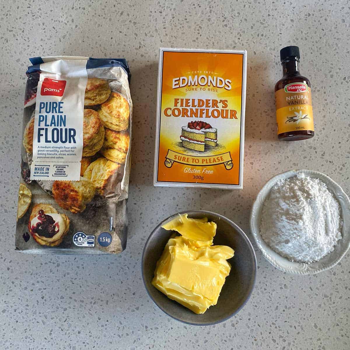 Ingredients on a countertop include plain flour, cornflour, vanilla extract, baking powder in a bowl, and a bowl of butter.
