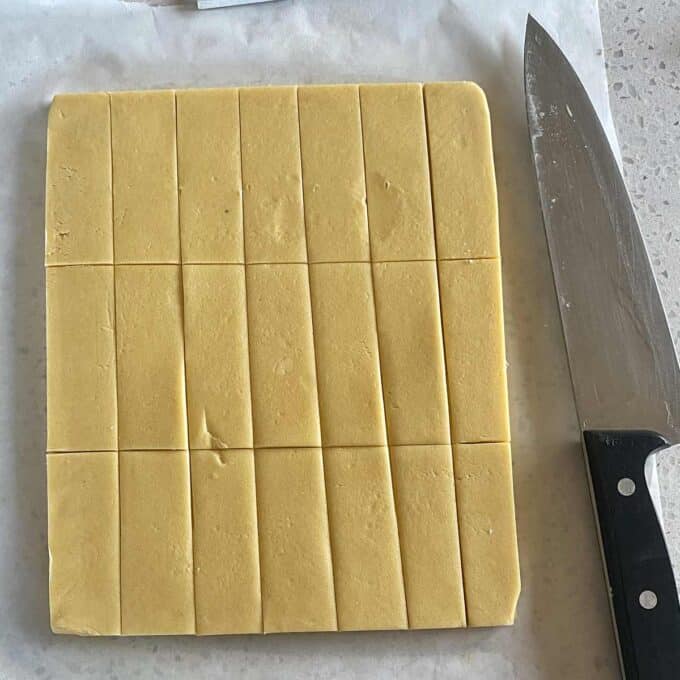 A square block of dough, scored into smaller rectangular sections, lies on parchment paper beside a large kitchen knife.