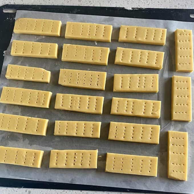 Rectangular pieces of uncooked shortbread dough with fork holes, arranged in rows on a parchment-lined baking tray.