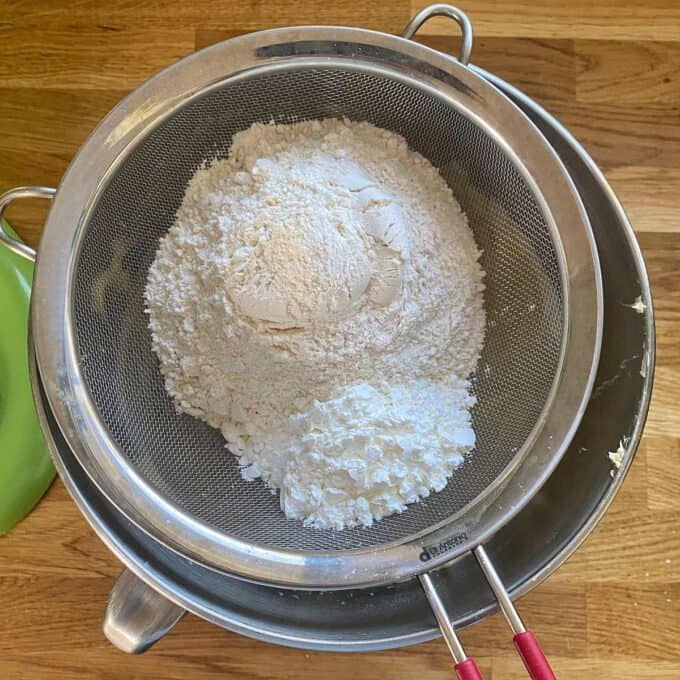 Flour being sifted through a metal sieve into a bowl on a wooden countertop.