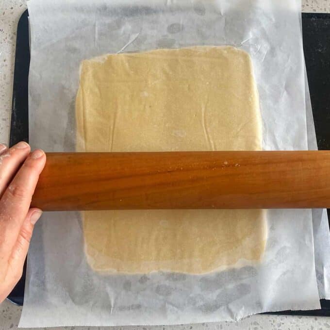 Hand using a rolling pin to flatten dough between two sheets of parchment paper on a countertop.
