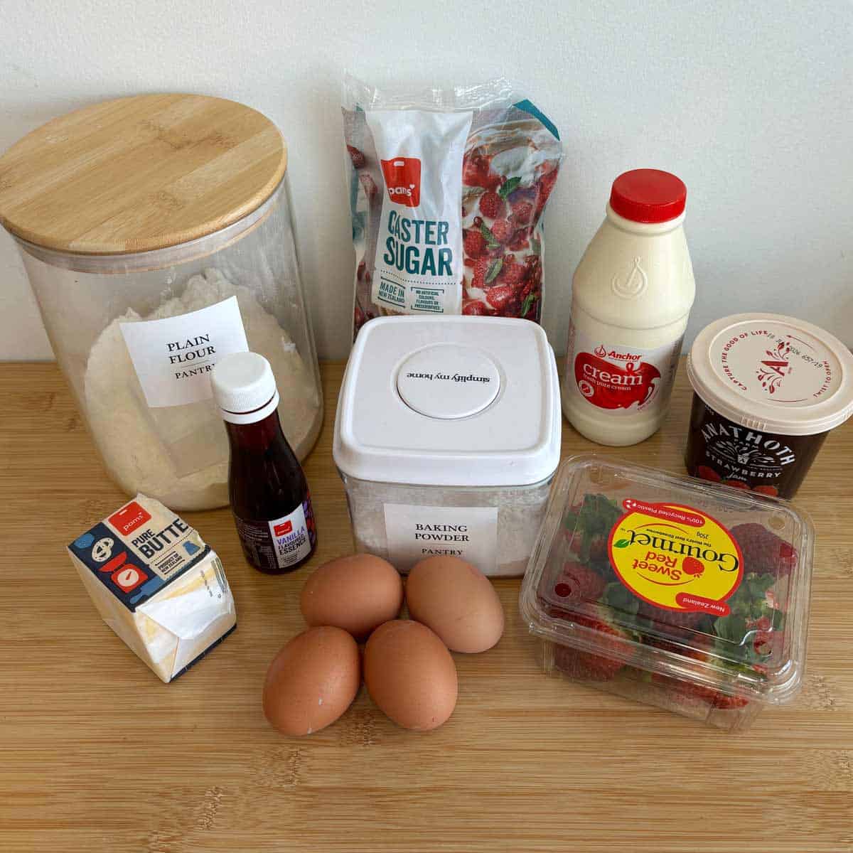 Ingredients for baking on a countertop: flour, caster sugar, baking powder, butter, vanilla extract, eggs, cream, yogurt, and strawberries.