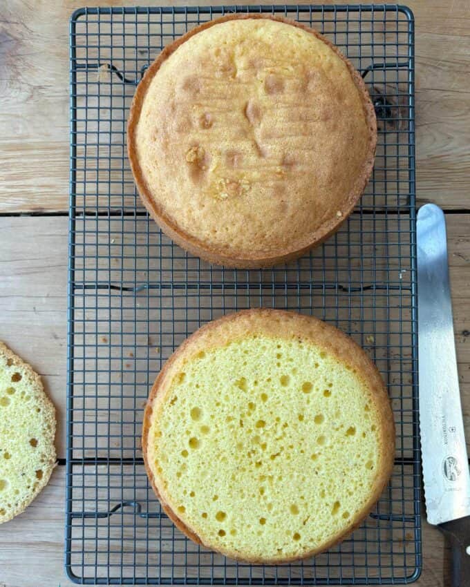 Two round sponge cakes cool on a wire rack placed on a wooden surface. A cake knife and a trimmed cake piece are visible beside the rack.