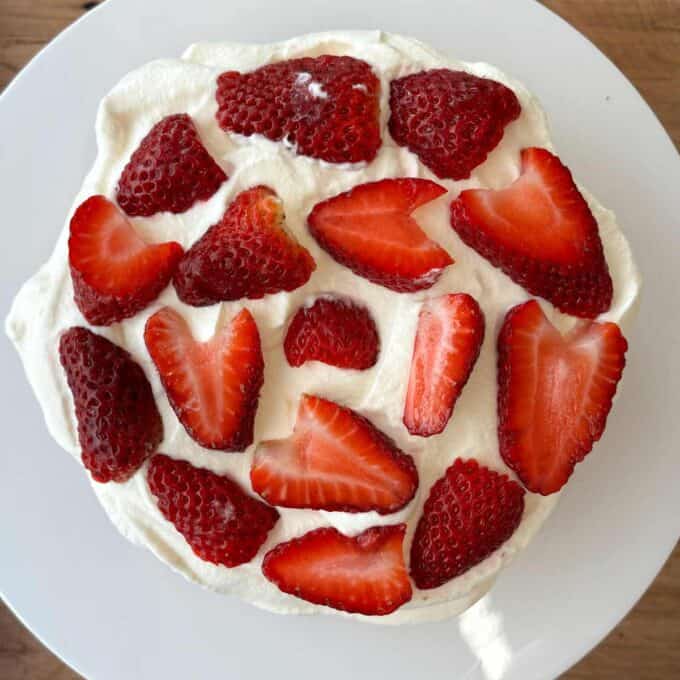 A round cake with whipped cream is topped with evenly spaced sliced strawberries, placed on a white plate.