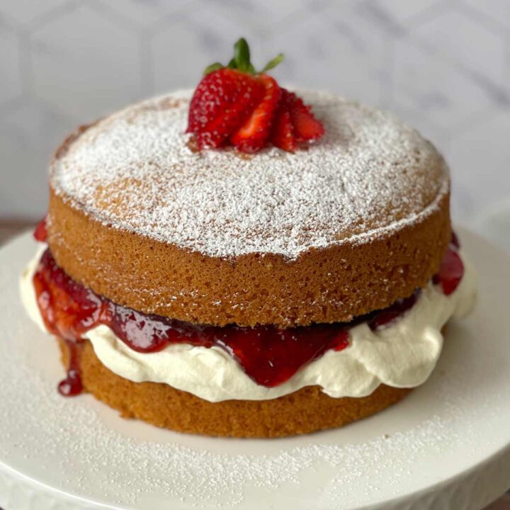 Two-layer sponge cake with cream and strawberry filling, topped with powdered sugar and sliced strawberries, on a white surface.
