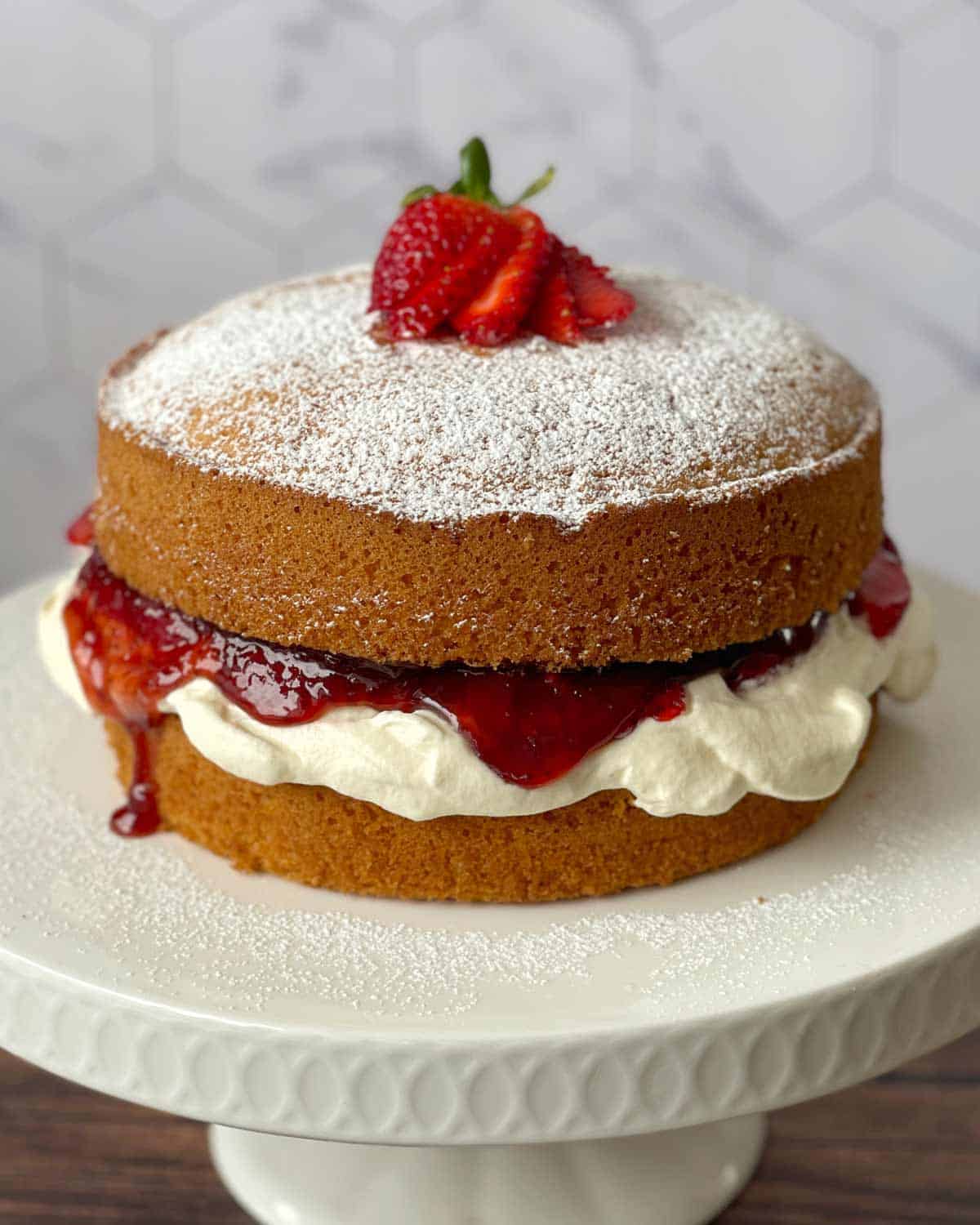 Victoria Sponge Cake Two-layer sponge cake on a white stand, filled with cream and strawberry jam, topped with powdered sugar and sliced strawberries.