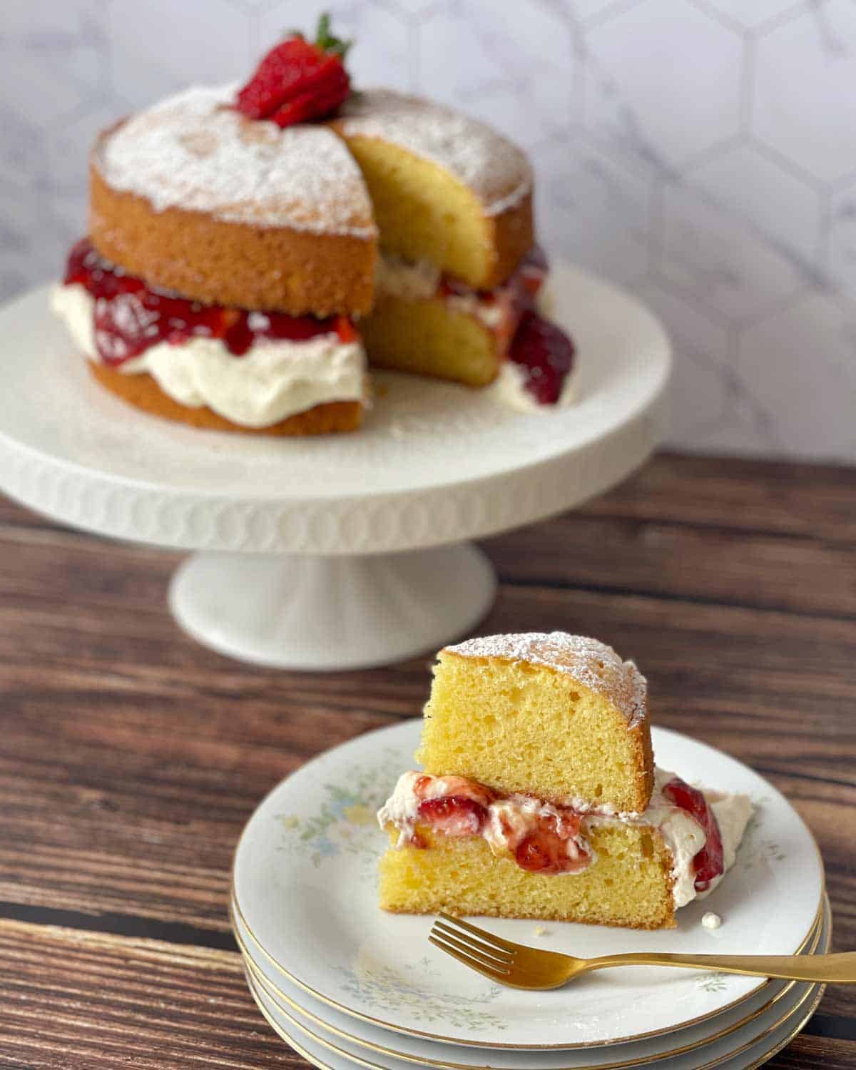 A slice of layered sponge cake with cream and strawberries on a plate, with the remaining cake on a stand in the background.